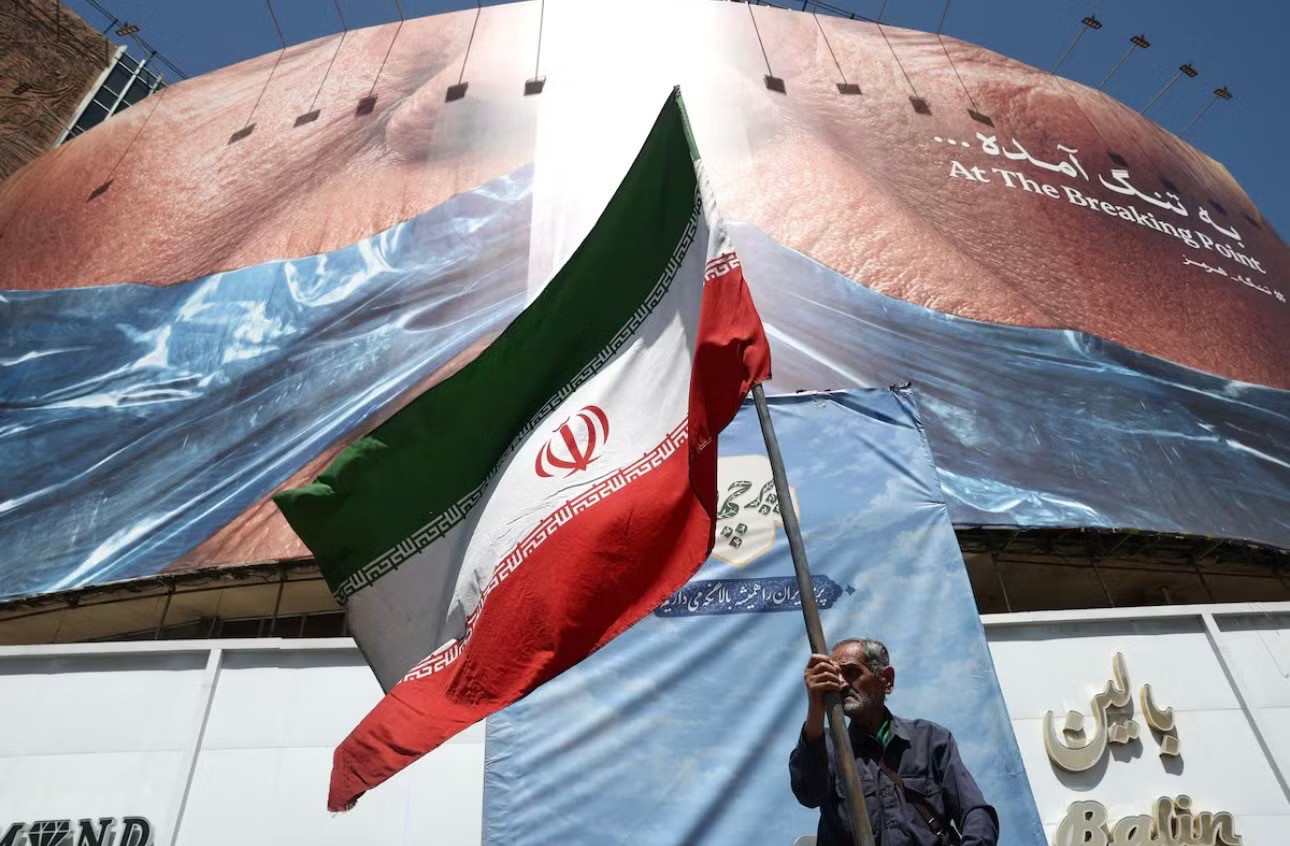 Một người giương cờ Iran gần tấm biểu ngữ mô tả Tổng thống Mỹ Donald Trump và eo biển Hormuz ở trung tâm thủ đô Tehran, ngày 30/5. (Ảnh: Reuters) Mỹ ra đòn với sàn tiền của gia tộc quyền lực bậc nhất Iran - Ảnh 1.