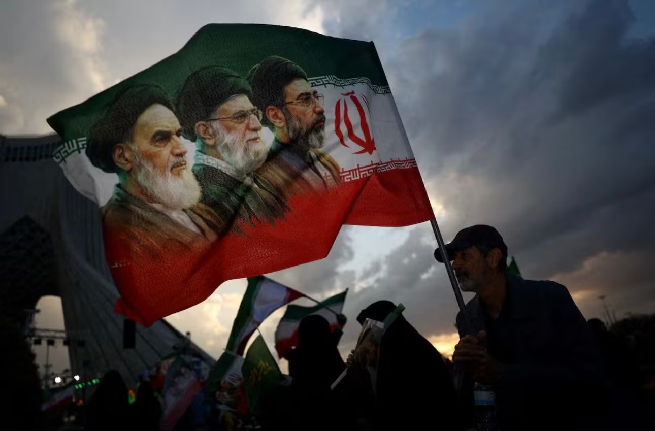 Một người dân Iran giương cờ in chân dung các lãnh tụ tối cao Iran. (Ảnh: AP) Iran chọn ngày tổ chức tang lễ cố Đại giáo chủ Ali Khamenei - Ảnh 1.