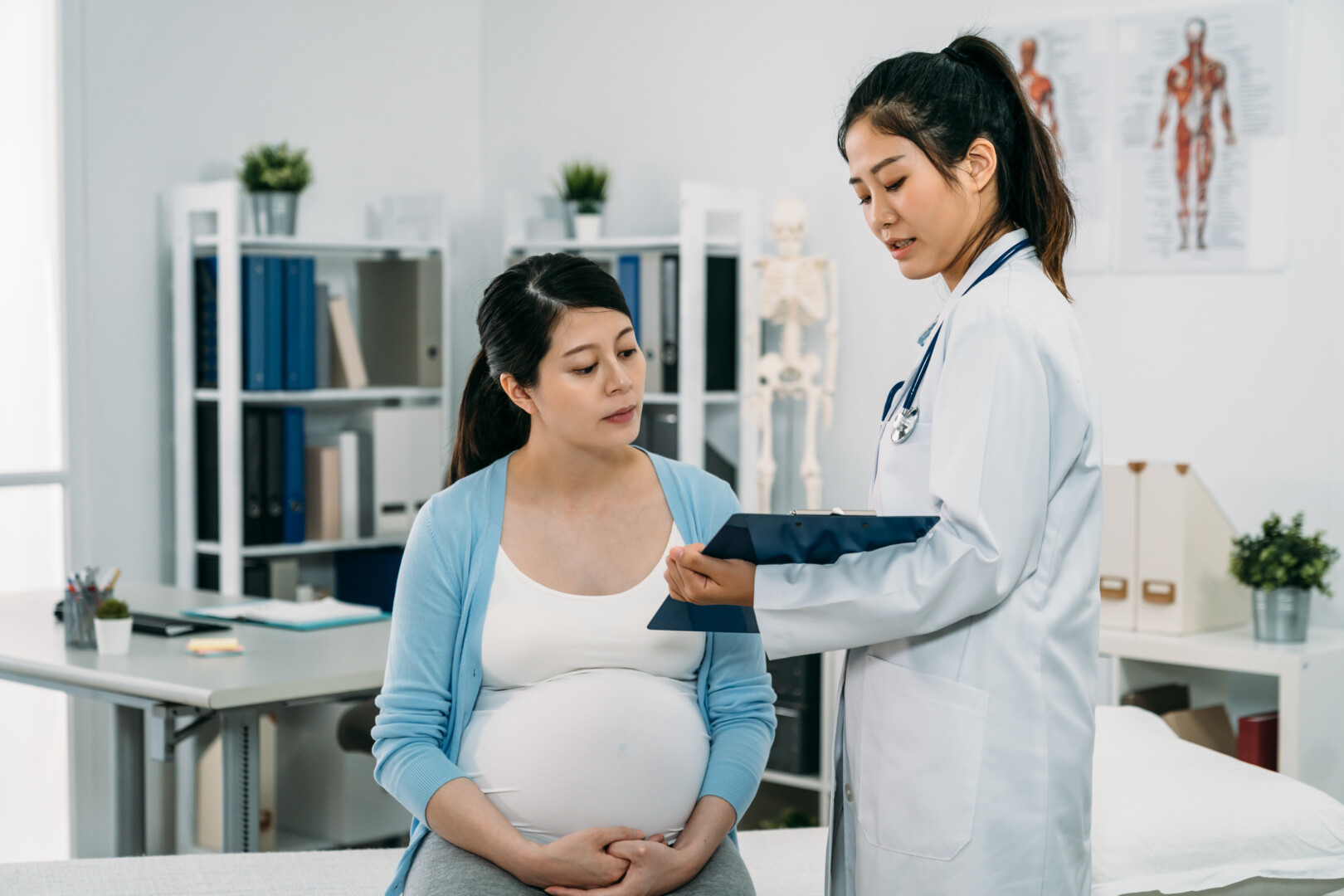 asian-female-physician-standing-with-clipboard-is-explaining-diagnosis-her-expectant-patient-who-s-sitting-treatment-bed-hospital.jpg